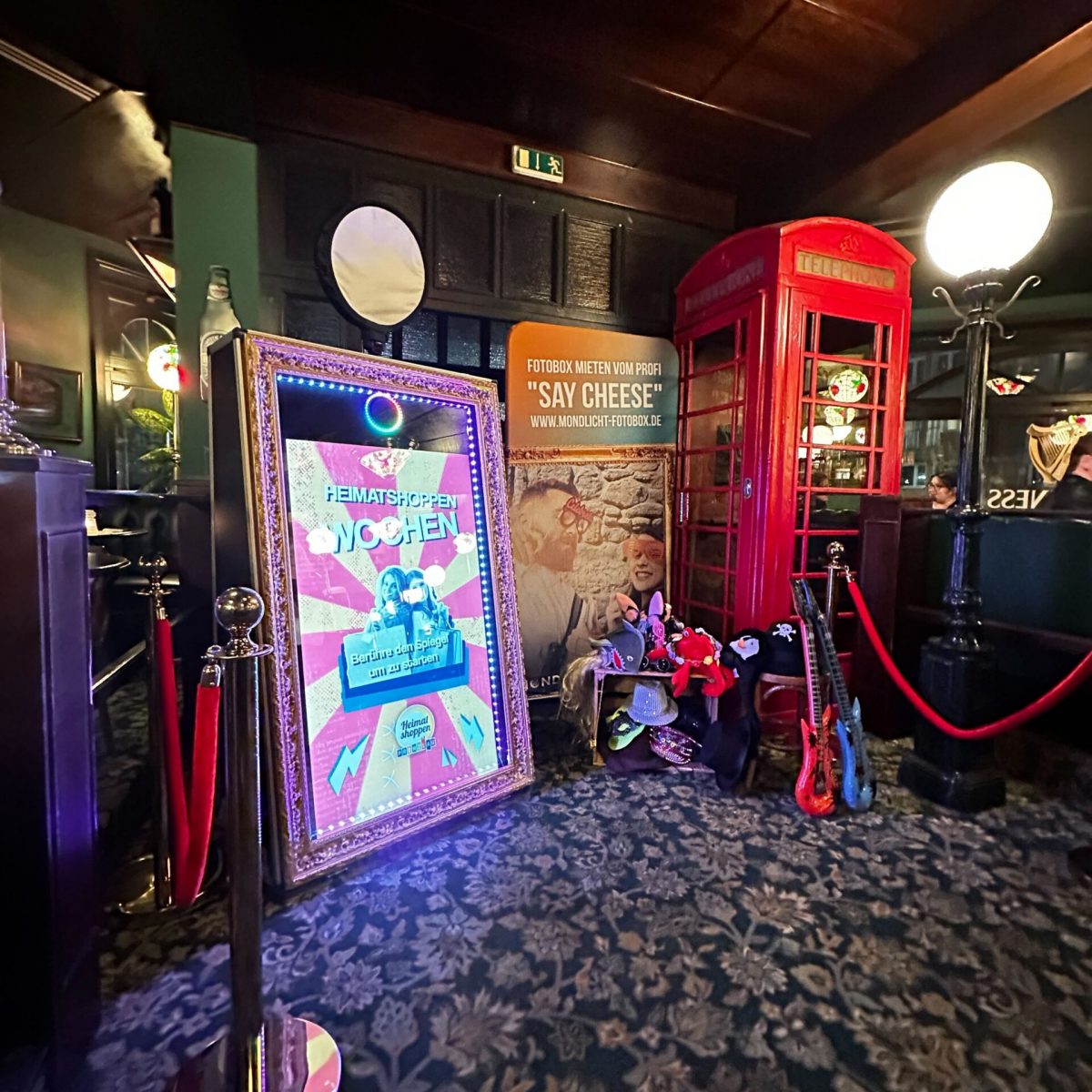 Ein Magic-Mirror-Fotospiegel mit LED-Lichtern steht in einem gemütlichen Raum mit einer roten Telefonzelle im Hintergrund. Ein Banner wirbt mit der Aufschrift 'Fotobox mieten vom Profi - Say Cheese'. Verschiedene Accessoires wie Hüte und Spielzeug-Gitarren sind aufgestellt.