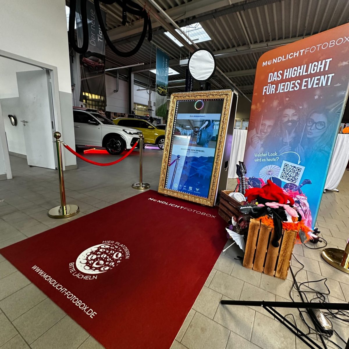 Ein Magic-Mirror-Fotospiegel mit einem roten Teppich und goldenen Absperrbändern steht in einem hellen Autohaus. Ein Banner im Hintergrund trägt die Aufschrift 'Das Highlight für jedes Event'. Daneben liegen eine Holzkiste mit bunten Accessoires und ein QR-Code-Banner.
