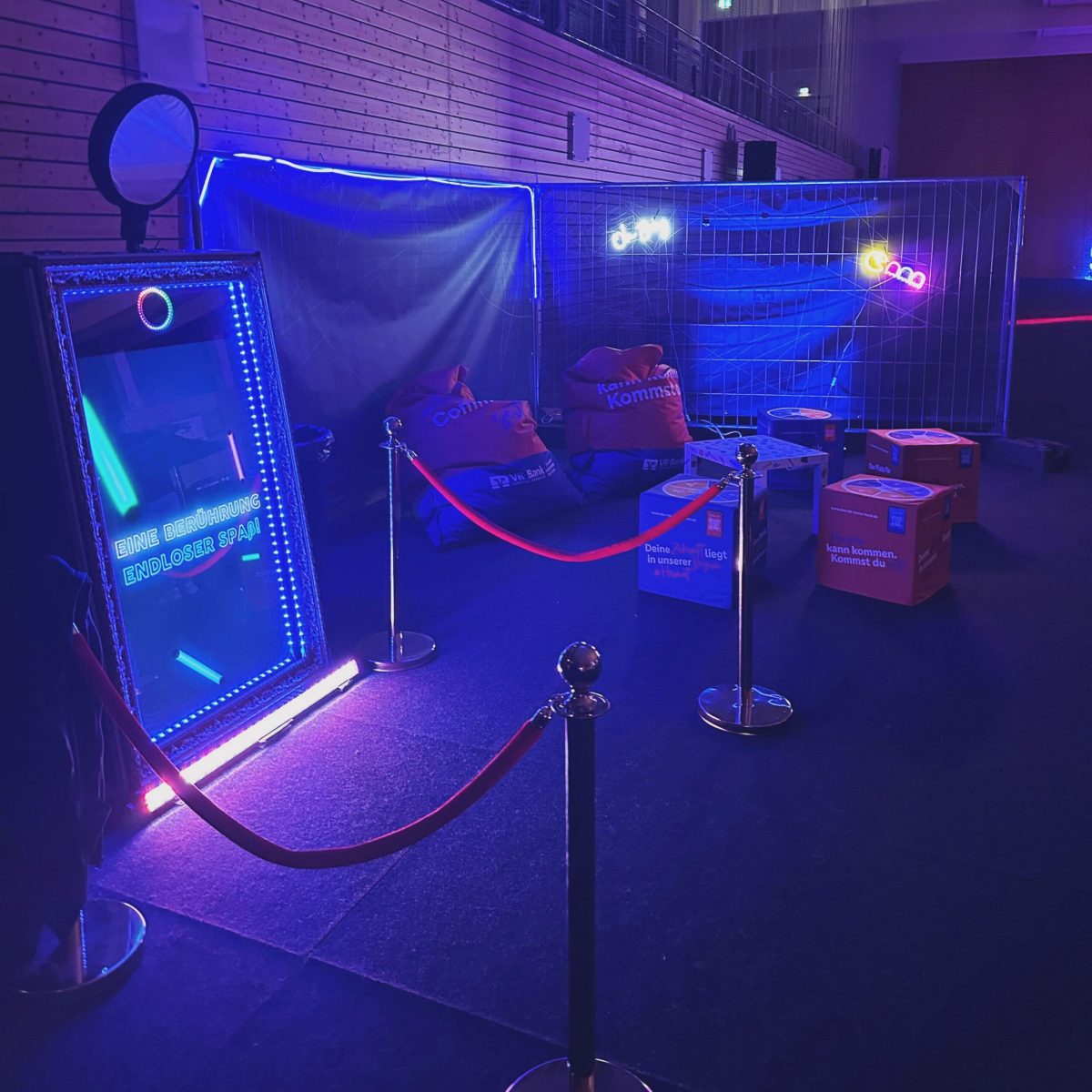 Ein Magic-Mirror-Fotospiegel in einer dunklen Halle, beleuchtet von bunten LED-Lichtern und umgeben von roten Absperrbändern. Im Hintergrund sind Sitzsäcke, ein Zaun und leuchtende Schilder zu sehen. Auf dem Spiegel steht der Text: 'Eine Berührung - endloser Spaß'.